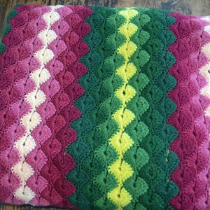 May include: A crocheted blanket with a striped pattern in shades of pink, green, and yellow. The blanket is made with a textured stitch that creates a raised, puffy effect.