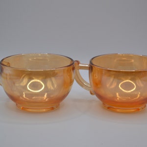 May include: Two amber glass cups with handles. The cups are iridescent and have a rounded shape.
