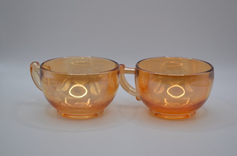 May include: Two amber glass cups with handles. The cups are iridescent and have a rounded shape.