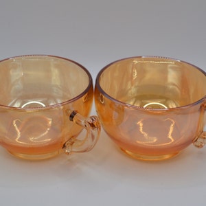 May include: Two iridescent orange glass teacups with handles. The cups have a rounded shape and a smooth, glossy finish.