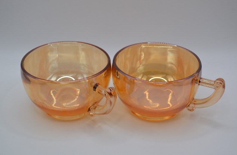 May include: Two iridescent orange glass teacups with handles. The cups have a rounded shape and a smooth, glossy finish.