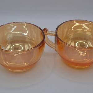 May include: Two amber glass cups with handles. The cups are iridescent and have a rounded shape.