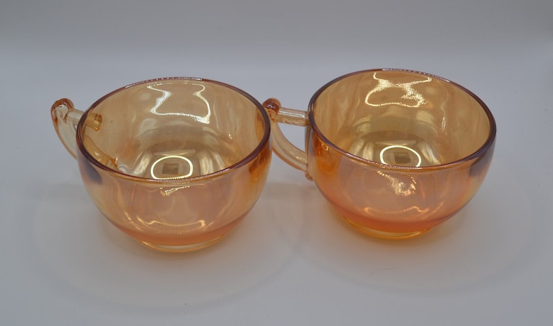 May include: Two amber glass cups with handles. The cups are iridescent and have a rounded shape.