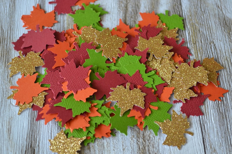May include: A collection of colorful, die-cut paper leaves in shades of orange, red, green, and gold glitter. The leaves are scattered, creating a festive autumn or fall theme. Ideal for crafting or seasonal decor.