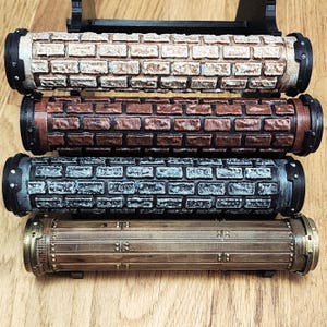 May include: Four cylindrical objects with textured surfaces. The top three have a brick pattern in beige, brown, and gray. The bottom one is bronze-colored with rivets and horizontal lines. All have black end caps and are displayed on a wooden surface.