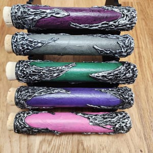 May include: A collection of five decorative bottle stoppers. Each stopper features a cylindrical body in shades of purple, gray, green, and pink, with a cork base. The stoppers are adorned with a silver, textured design.