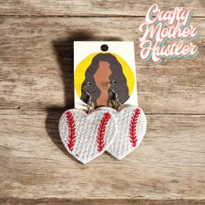 May include: A pair of heart-shaped earrings with a white and red baseball design. The earrings are hanging from silver hooks.