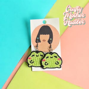 May include: A pair of green frog-shaped earrings with pink cheeks. The earrings are hanging from a white card with a drawing of a woman with black hair.