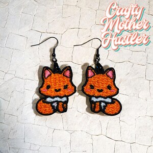 May include: A pair of orange fox-shaped dangle earrings with black hooks. The foxes have white fur around their necks and pink ears.