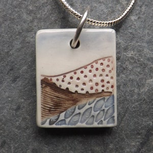 Peut inclure: Un collier avec un pendentif en céramique blanche avec un motif abstrait brun, rouge et bleu. Le pendentif est de forme rectangulaire et est sur une chaîne argentée.