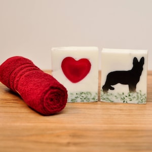 Puede incluir: Dos barras de jabón blancas con un corazón rojo y una silueta negra de un perro pastor alemán. Las barras de jabón están sobre una superficie de madera con una toalla roja.