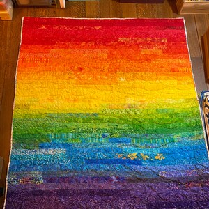 May include: A rainbow-colored quilt with a patchwork design. The quilt is made up of different shades of red, orange, yellow, green, blue, indigo, and violet. The quilt is made of fabric scraps sewn together in a horizontal pattern.