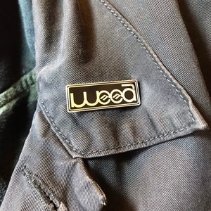 May include: A silver metal pin with a black rectangular background and the word "weed" in white letters. The letters are stylized with a wavy design.