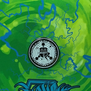 May include: A black and white enamel pin with a circular design. The pin features a stylized figure sitting in a chair with a circle around it. The background is a green and blue swirl pattern.