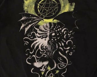 Puede incluir: Camiseta negra con un estampado gráfico de una criatura con tentáculos y alas, un pentagrama y una luna amarilla.