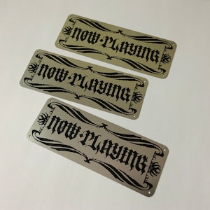 Könnte beinhalten: Drei silberne Metallschilder mit schwarzer Beschriftung "Now Playing". Die Schilder sind rechteckig und haben abgerundete Ecken.