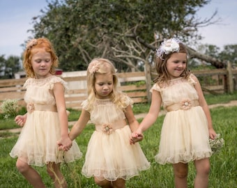 May include: Three young girls in matching cream-colored dresses with floral accents and lace details, holding hands. They are standing in a grassy field near a wooden fence, with flowers in their hands.