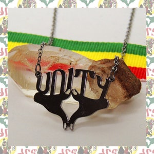 May include: A silver-toned necklace spelling "UNITY" in a stylized font. The pendant features a design of two hands clasped together. The necklace rests on a rough, light-brown stone, with a ribbon of green, yellow, and red in the background.