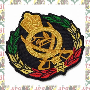 Puede incluir: Parche bordado con un diseño de león y espada dorados rodeado de hojas verdes y rojas. El parche está sobre un fondo negro.