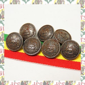 Puede incluir: Seis botones antiguos de bronce con un diseño de león y escritura etíope. Los botones están dispuestos en una cinta de rayas rojas, amarillas y verdes.