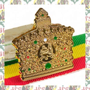 Puede incluir: Un broche dorado en forma de corona ornamentada, con detalles verdes y rojos. El broche presenta un diseño detallado y se muestra sobre un fondo a rayas rojo, amarillo y verde, lo que sugiere un tema cultural o histórico.