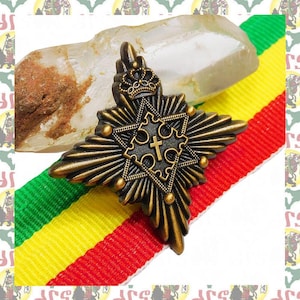Puede incluir: Un pin de metal dorado en forma de estrella con una cruz en el centro. El pin está en una cinta de rayas rojas, amarillas y verdes.
