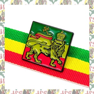 May include: A green, yellow, and red enamel pin featuring a lion holding a spear and a flag. The lion is standing on a red background with a green and yellow flag behind it.