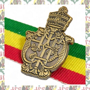 May include: A gold-colored metal pin with a crown and intricate design. The pin is on a red, yellow, and green striped ribbon.