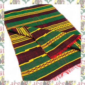May include: A rectangular textile with a striped pattern in green, yellow, and maroon. The fabric has a fringed edge in red. The design includes horizontal stripes and decorative woven sections. The textile is folded over, displaying the pattern.