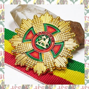 Op de afbeelding: Een gouden stervormige medaille met een rood en groen kruis in het midden. Het kruis heeft een groene cirkel met een leeuw in het midden. De medaille ligt op een rood, geel en groen lint.