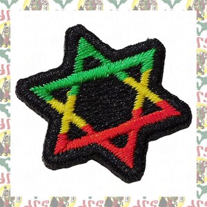 May include: Embroidered patch in the shape of a six-pointed star. The star features green, yellow, and red stitching on a black background. The patch has a textured appearance and is likely designed to be sewn or ironed onto fabric.