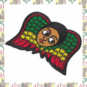 May include: Embroidered patch featuring a stylized angel with wings in red, yellow, and green. The angel has a brown face with large eyes and a serious expression.