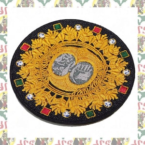 May include: A round, black embroidered patch with a gold design. The center of the patch features two silver hands clasped together. The gold design is a circular pattern of leaves and diamonds.