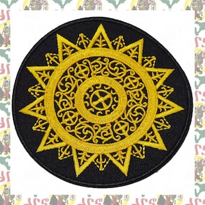 May include: A round black patch with a gold embroidered design. The design features a large, intricate sun with a cross in the center.