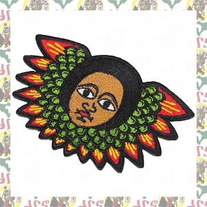 Puede incluir: Parche bordado con un rostro estilizado con cabello negro, piel marrón y ojos grandes. El rostro está rodeado por un diseño floral verde y amarillo.