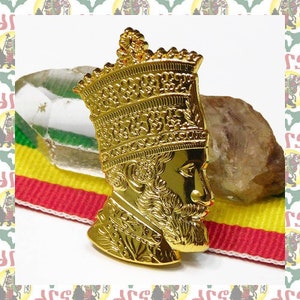 Puede incluir: Un pin de metal dorado que representa el perfil de una persona con una corona. El pin está detallado con diseños intrincados y probablemente representa una figura histórica.