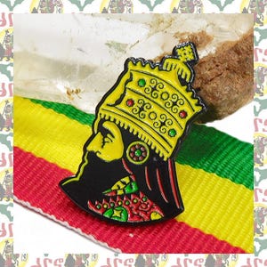 Puede incluir: Un pin de metal esmaltado que muestra la silueta de perfil de una figura con una corona amarilla con detalles en rojo y verde. La figura tiene el rostro y la barba negros, con patrones decorativos en rojo, amarillo y verde en el cuello.