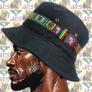 Könnte beinhalten: Schwarzer Fischerhut mit einem farbenfrohen Band mit Sound-System-Grafiken und dem Text "SOUND SYSTEM". Der Hut hat zwei Messingösen und eine breite Krempe. Der Hintergrund hat ein sich wiederholendes Muster.