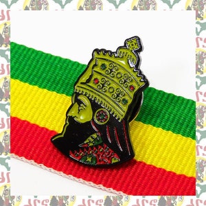 May include: A gold and green enamel pin depicting a king wearing a crown and a black robe. The pin is on a red, yellow, and green striped fabric.
