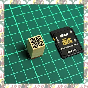May include: A wooden stamp with a black and white geometric design on a green cutting mat next to an 8GB SDHC memory card. The memory card is black with gold lettering and says "SDHC Memory Card" and "JAPAN".