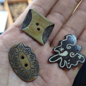 May include: Three decorative wooden buttons in various shapes. One is a light brown square with dark brown accents and two holes. Another is an oval with a floral design and two holes. The third is a dark brown tree-shaped button with white accents.