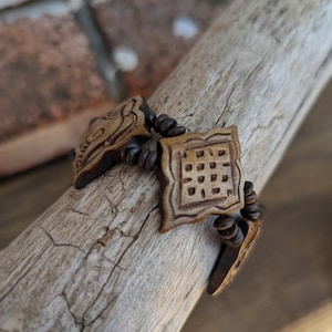 May include: A handcrafted wooden bracelet with intricate carvings. The bracelet features a central square design with a grid pattern, flanked by carved decorative elements and connected by dark brown beads. The bracelet is displayed on a piece of driftwood.