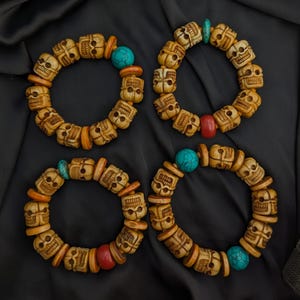 May include: Four circular bracelets composed of carved beige skull-shaped beads interspersed with flat orange spacers, smooth red beads, and round turquoise-colored beads with dark veining, arranged in a grid pattern on a dark fabric surface.