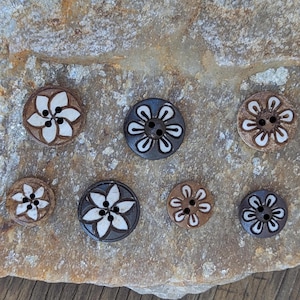 May include: A collection of seven decorative wooden buttons with floral designs. The buttons feature a variety of colors, including brown, white, and dark gray. Each button has two holes for sewing and a unique flower pattern.