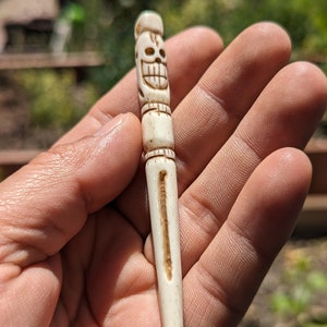 May include: A white bone carving of a skull with a toothy grin. The carving is detailed with lines and a small hole at the top. The carving is held in a hand.