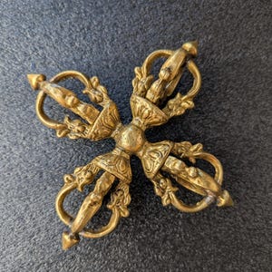 May include: A detailed, antique-style brass vajra, a ritual object with a cross-shaped design. The vajra features intricate floral and geometric patterns, with pointed ends and curved handles. The object is displayed on a dark gray surface.