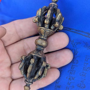 May include: A bronze-colored metal vajra ritual object featuring a central grip and two symmetrical, multi-pronged ends. The surface displays a weathered, dark patina with highlights on the raised edges of the intricate, geometric metalwork.