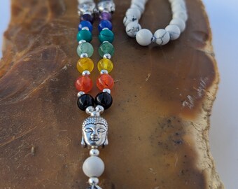 May include: A multi-colored beaded necklace with a silver Buddha charm and a white stone bead. The necklace is on a brown background.