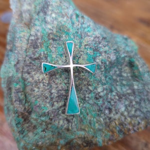 May include: A silver cross pendant with turquoise inlay. The cross is set against a green and grey background.