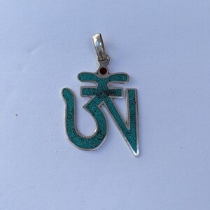 May include: A silver pendant with a turquoise Om symbol and a red stone in the center.
